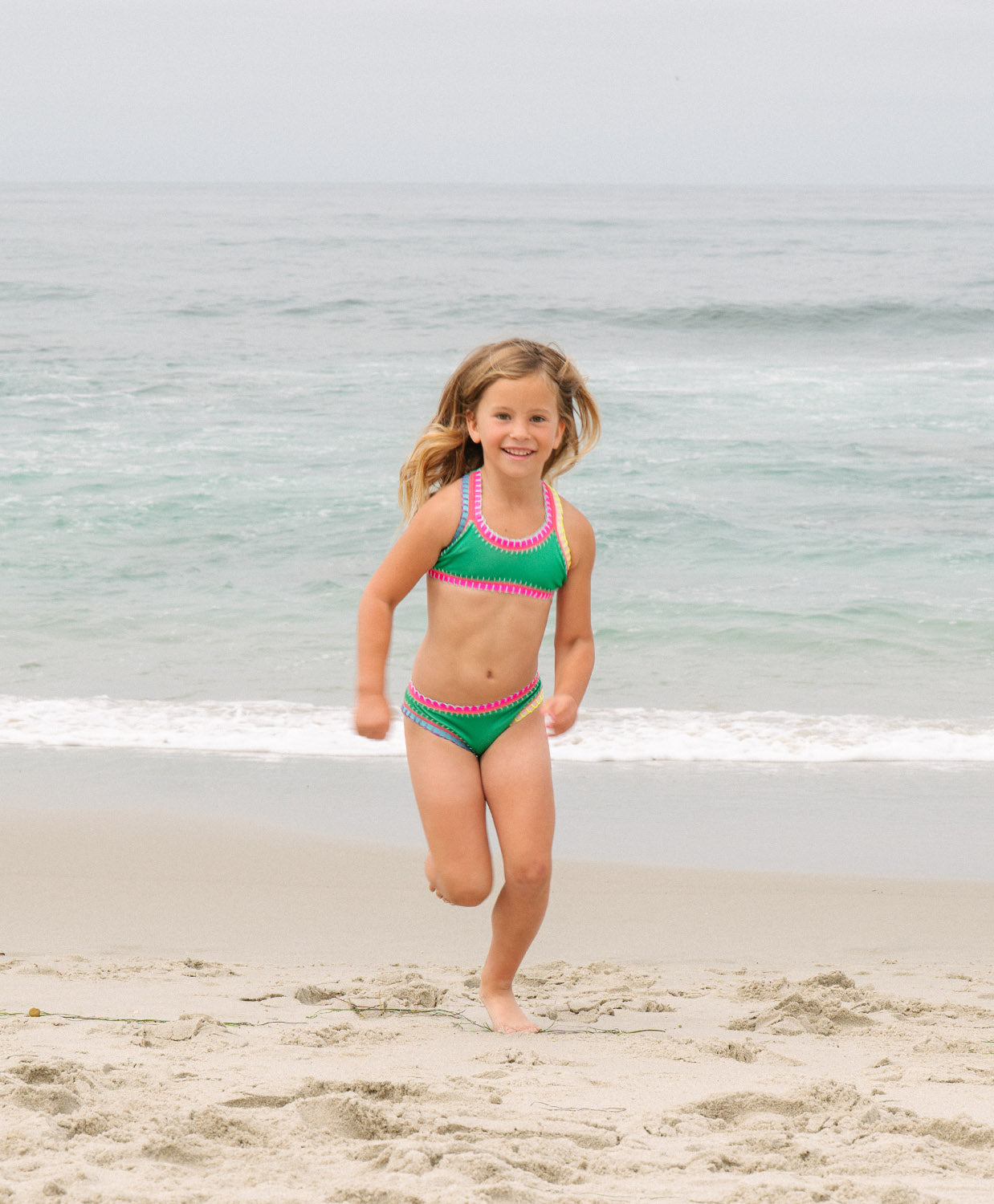 Young girl in a green bikini with pink trim standing on a beach.