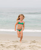 Young girl in a green bikini with pink trim standing on a beach.