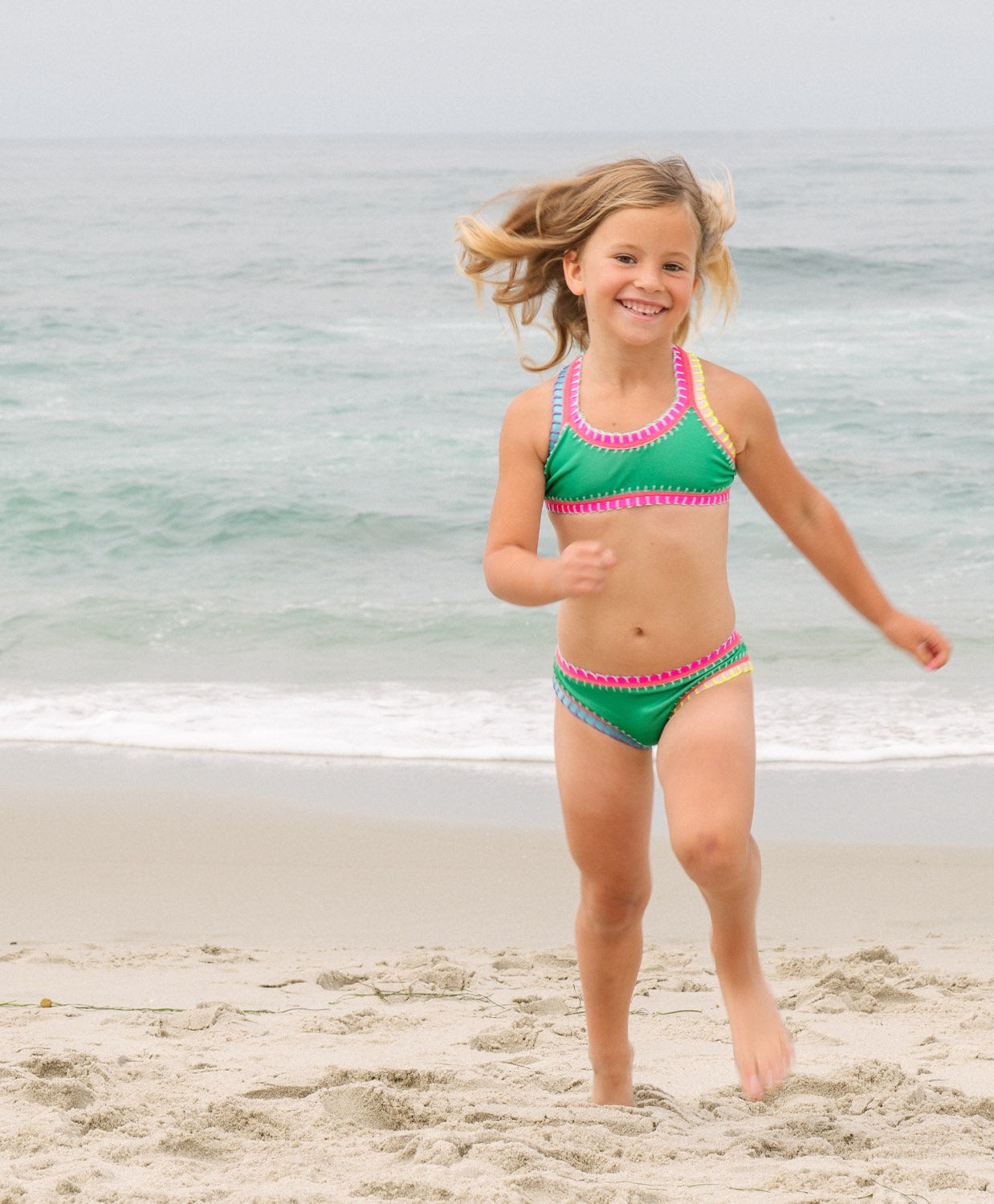 Young girl in a green and pink swimsuit running on a beach with ocean in the background
