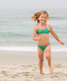 Young girl in a green and pink swimsuit running on a beach with ocean in the background