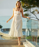 A blonde woman wearing a midi length white dress standing outside near a glass wall. 