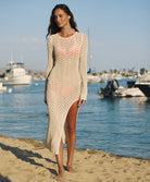 A woman wearing a long sleeve ivory dress standing on a beach near an ocean.