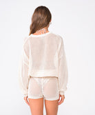A woman wearing an ivory knit sweater and matching shorts facing towards a white wall.