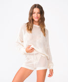 A woman wearing an ivory knit sweater and matching shorts standing against a white wall.