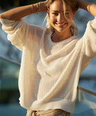 A blonde woman wearing an ivory knit long sleeve sweater leaning against a glass wall outside. 