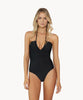 Blonde woman wearing a black one piece swimsuit standing in front of white wall.