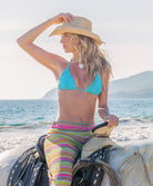 Woman in a blue bikini and colorful striped pants sitting on a horse by the ocean.