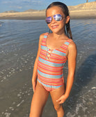 A young girl wearing a striped one piece swimsuit standing on a beach near the ocean. 