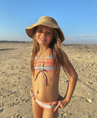 A young girl wearing a striped bikini and bucket hat standing on a beach.