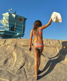 A young girl wearing a striped bikini and holding a bucket hat walking on the beach.