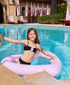 A brunette girl wearing a black bikini playing in a pool with a pink inflatable floaty. 