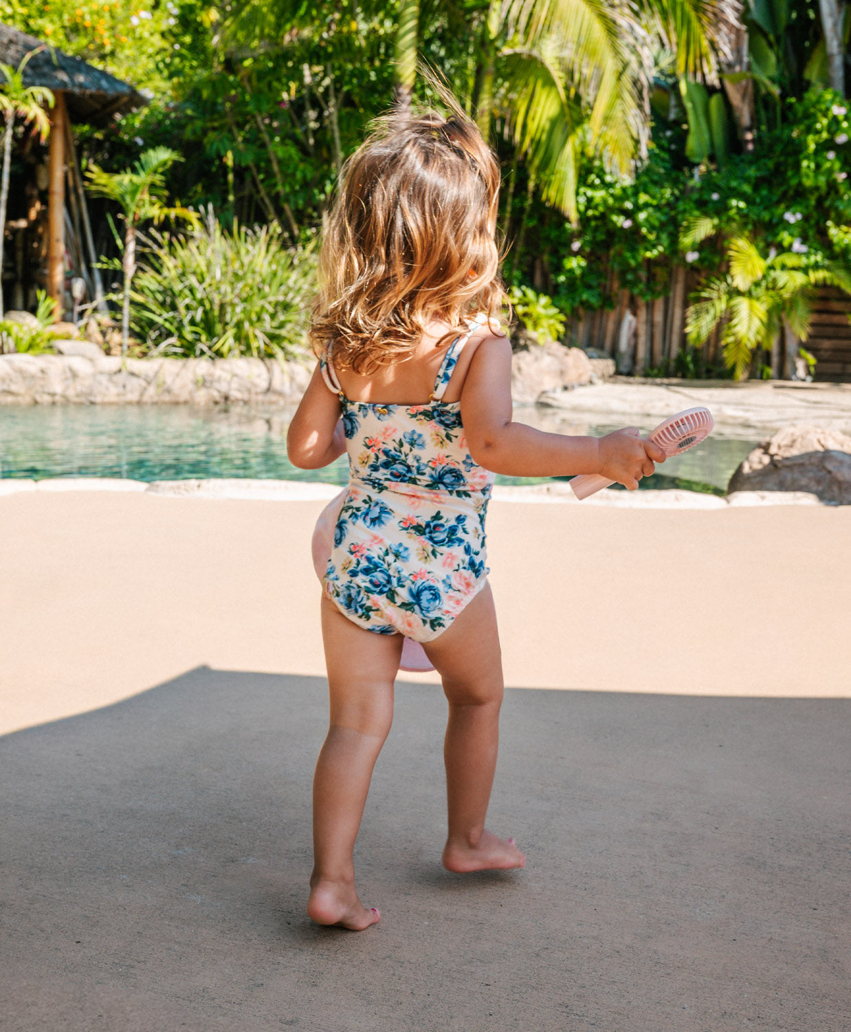 Child in a floral swimsuit standing by a pool with greenery in the background