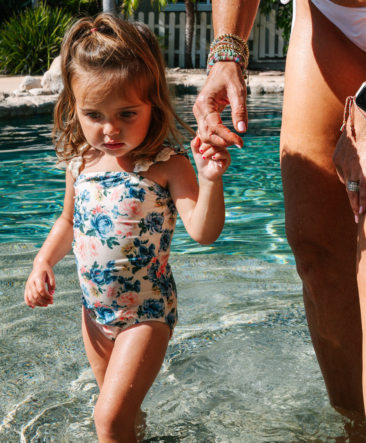Child in a floral swimsuit holding an adult's hand by a pool
