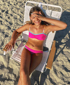 A young girl wearing a pink bikini with blue trim sitting on a chair on a beach.