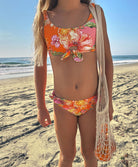 A young girl wearing a pink and orange bikini with a floral print standing on a beach.