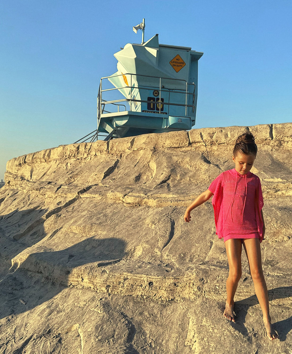 A young girl wearing a pink knit poncho walking on a beach near a lifeguard tower.