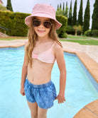 A brunette girl wearing a light pink bikini and denim shorts standing hear a pool. 