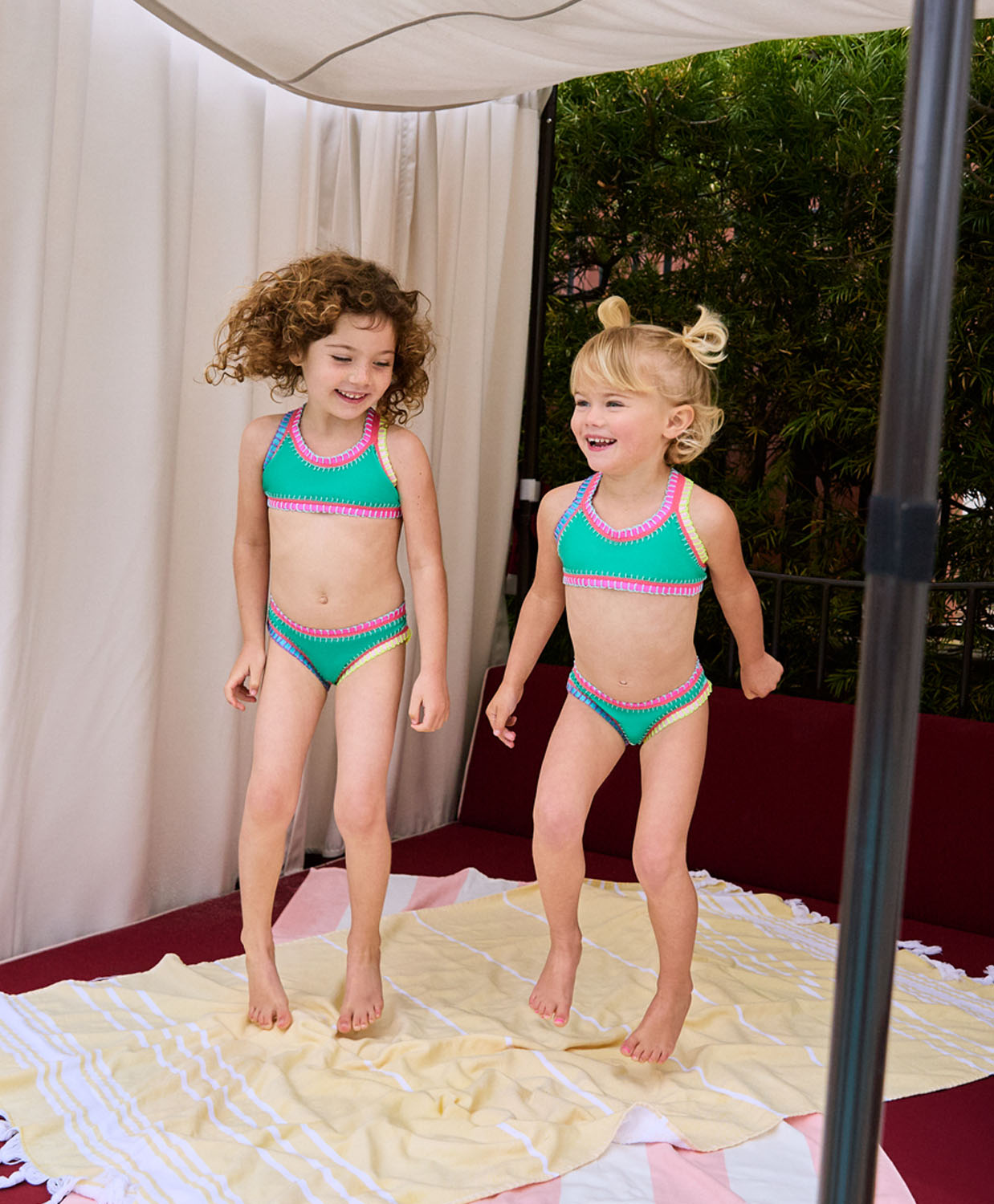 Two girls wearing green bikinis with colorful embroidery standing on a beach towel. 