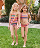 A blonde girl wearing a striped one piece swimsuit and a brunette girl wearing a striped bikini. Both girls are outside standing on grass. 