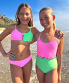 Two young girls standing on a beach near an ocean. One girl is wearing a pink and green bikini and the other girl is wearing a pink and green one piece.