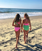 Two young girls standing on a beach near an ocean. One girl is wearing a pink and green bikini and the other girl is wearing a pink and green one piece.