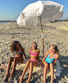 Three young girls sitting on a beach under an umbrella. One girl is wearing a turquoise bikini, another girl is wearing a hot pink bikini, and another girl is wearing a turquoise one piece.