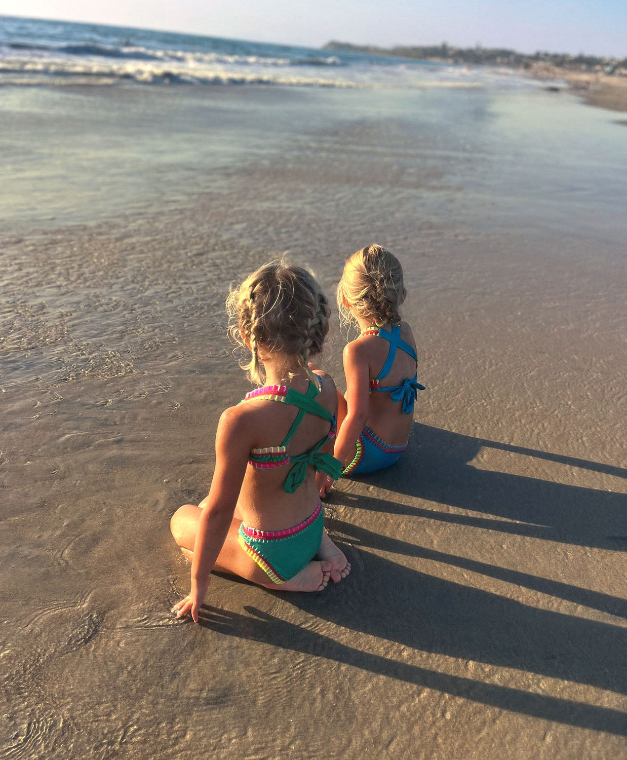 Two girls sitting on a beach near the ocean. One girl is wearing a green embroidered bikini and the other girl is wearing a turquoise embroidered bikini.