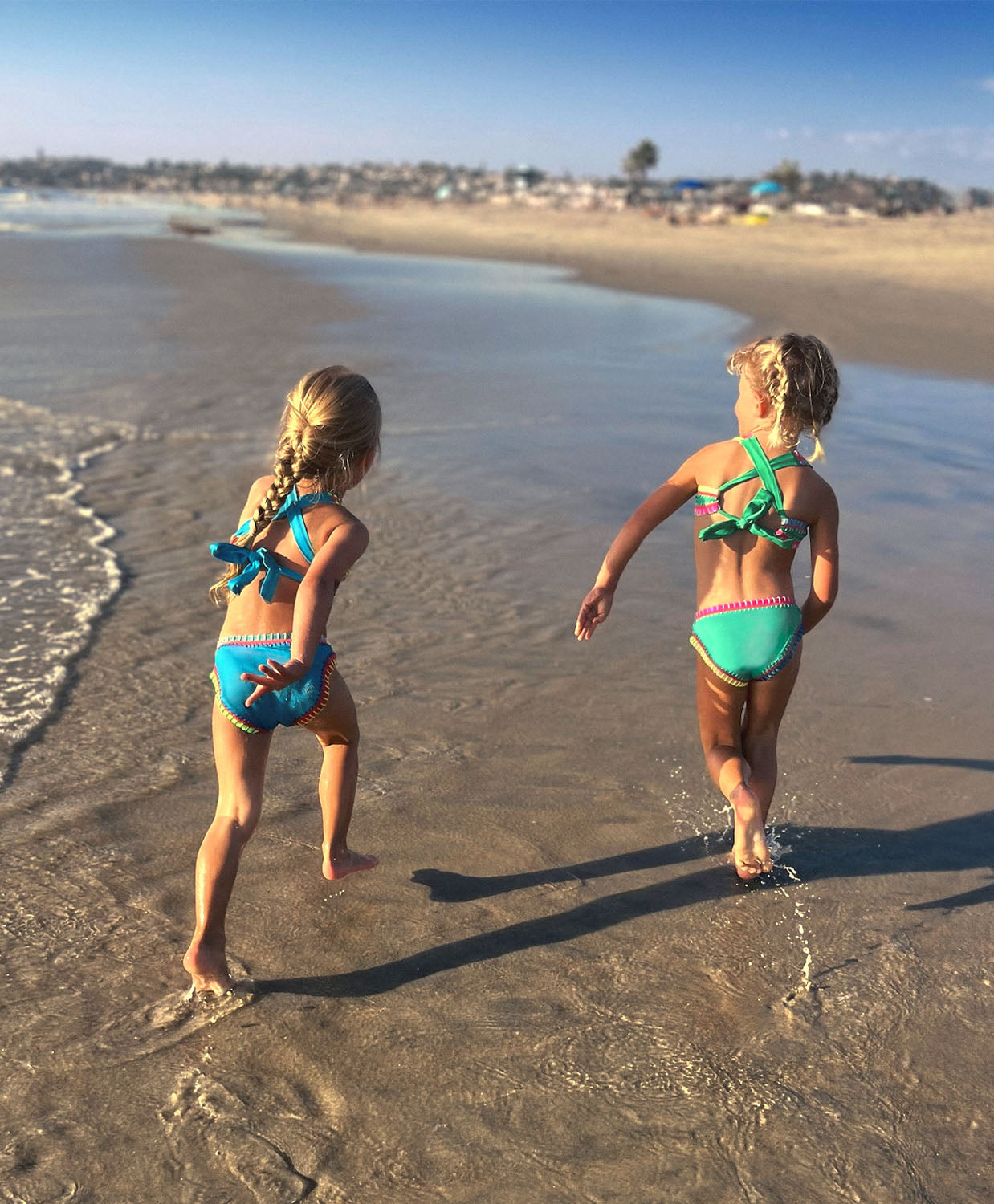 Two girls walking on a beach near the ocean. One girl is wearing a green embroidered bikini and the other girl is wearing a turquoise embroidered bikini.