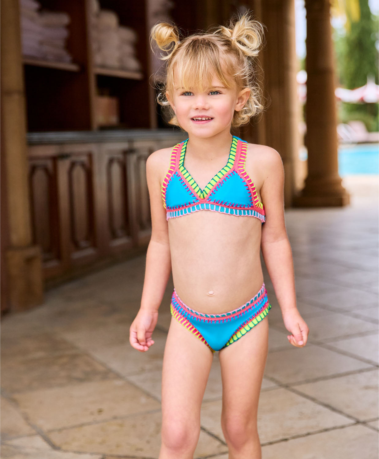 A blonde girl wearing a turquoise bikini with colorful embroidery standing near a pool. 