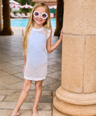 A blonde girl wearing a white knit cover up dress standing near a pool. 