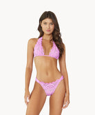 Brunette woman wearing a violet lace halter bikini stands in front of white wall.