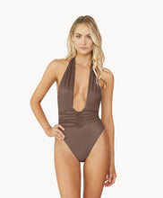 A blonde woman wearing a brown one piece bathing suit stands in front of a white wall. 