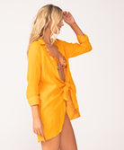 A woman wearing an orange bikini and orange long sleeve cover up standing against a white wall.