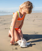 Child on a beach in an orange bikini with a large shell.