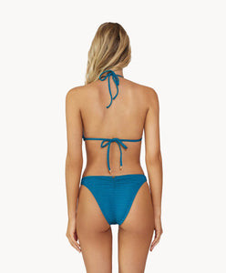 Blonde woman wearing a textured turquoise triangle shape bikini with gold details facing backwards towards a white wall.