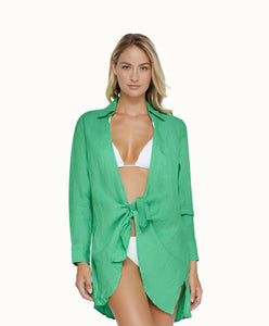 A blonde woman wearing a white bikini and a green long sleeve cover up against a white wall. 