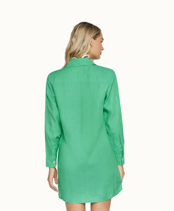 A blonde woman wearing a long sleeve green cover up facing towards a white wall. 