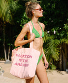 Woman in green bikini holding a pink bag with 'Vacation is the Answer' text, standing in a tropical setting.