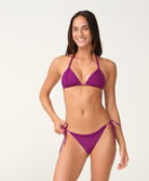 A woman modeling a purple bikini with textured crochet-style detailing on the cups and sides, featuring a halter top and side-tie bottoms, against a plain light background.