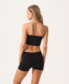 A woman wearing a black bandeau and matching shorts facing towards a white wall.