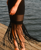 A close-up of a model wearing a black knit fringe skirt with a fishnet pattern, standing on a wooden dock with water in the background.