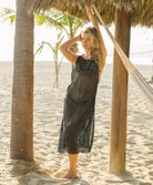 Woman in a black patterned dress standing on a sandy beach with palm trees and a hammock in the background.