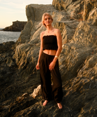 A woman stands barefoot on rocky coastal cliffs at sunset, wearing a black strapless cropped tube top and matching black wide-leg pants.
