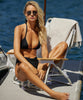 A blonde woman wearing a black bikini sitting on a chair near a body of water.