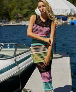 A blonde woman wearing a multi-colored floor length dress standing on a dock near a boat. 