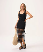 A woman wearing a short black slip dress under a black crochet dress standing against a white wall.