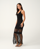 A woman modeling a black sleeveless crochet maxi dress with a fringed hem, photographed against a plain light gray studio background.