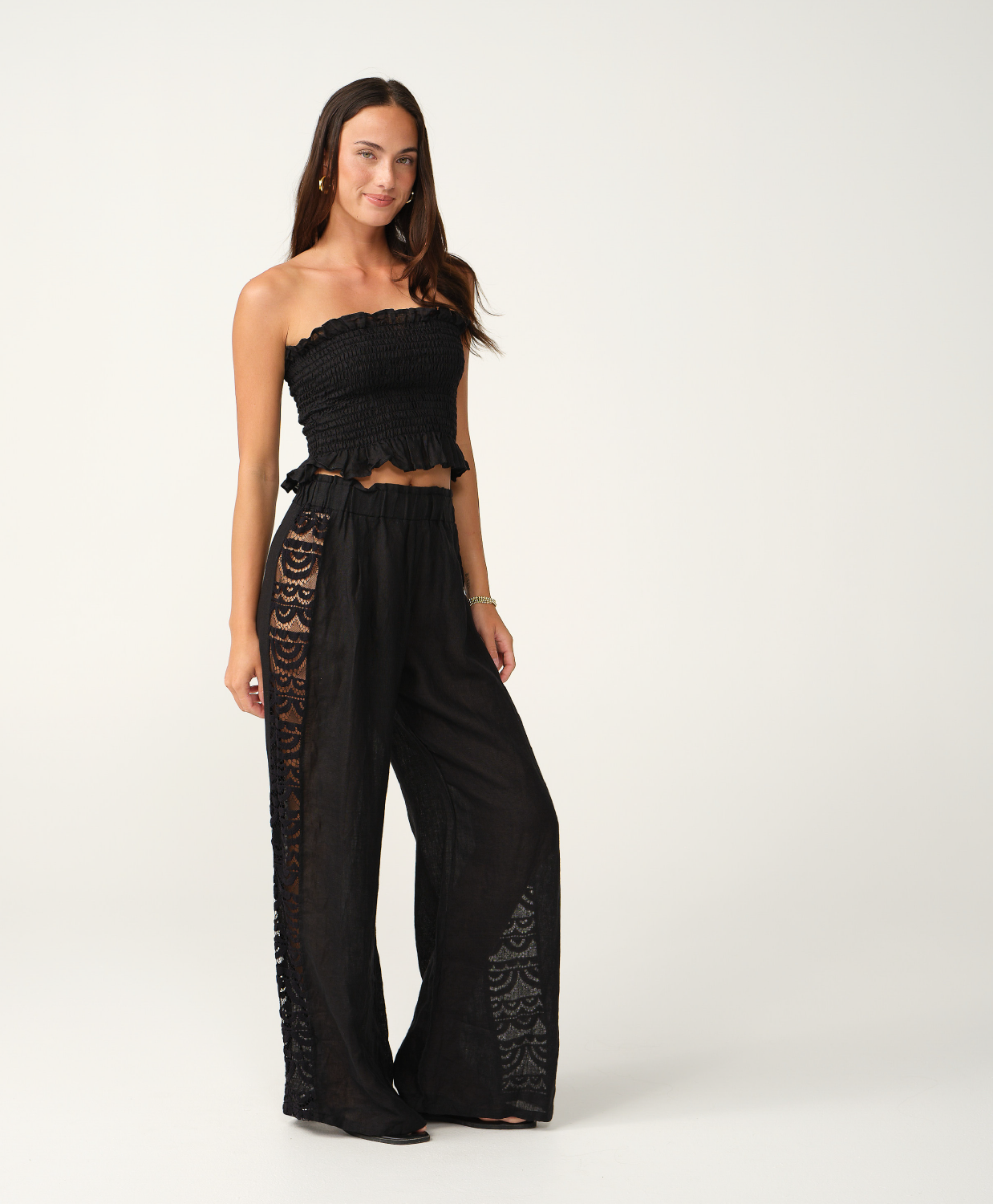 A woman modeling a black strapless cropped top and matching wide-leg pants with lace side panels, photographed against a clean off-white studio background.