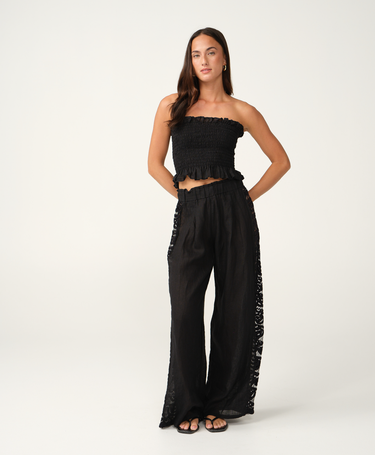 A woman modeling a black strapless cropped top and matching wide-leg pants with lace side panels, photographed against a clean off-white studio background.