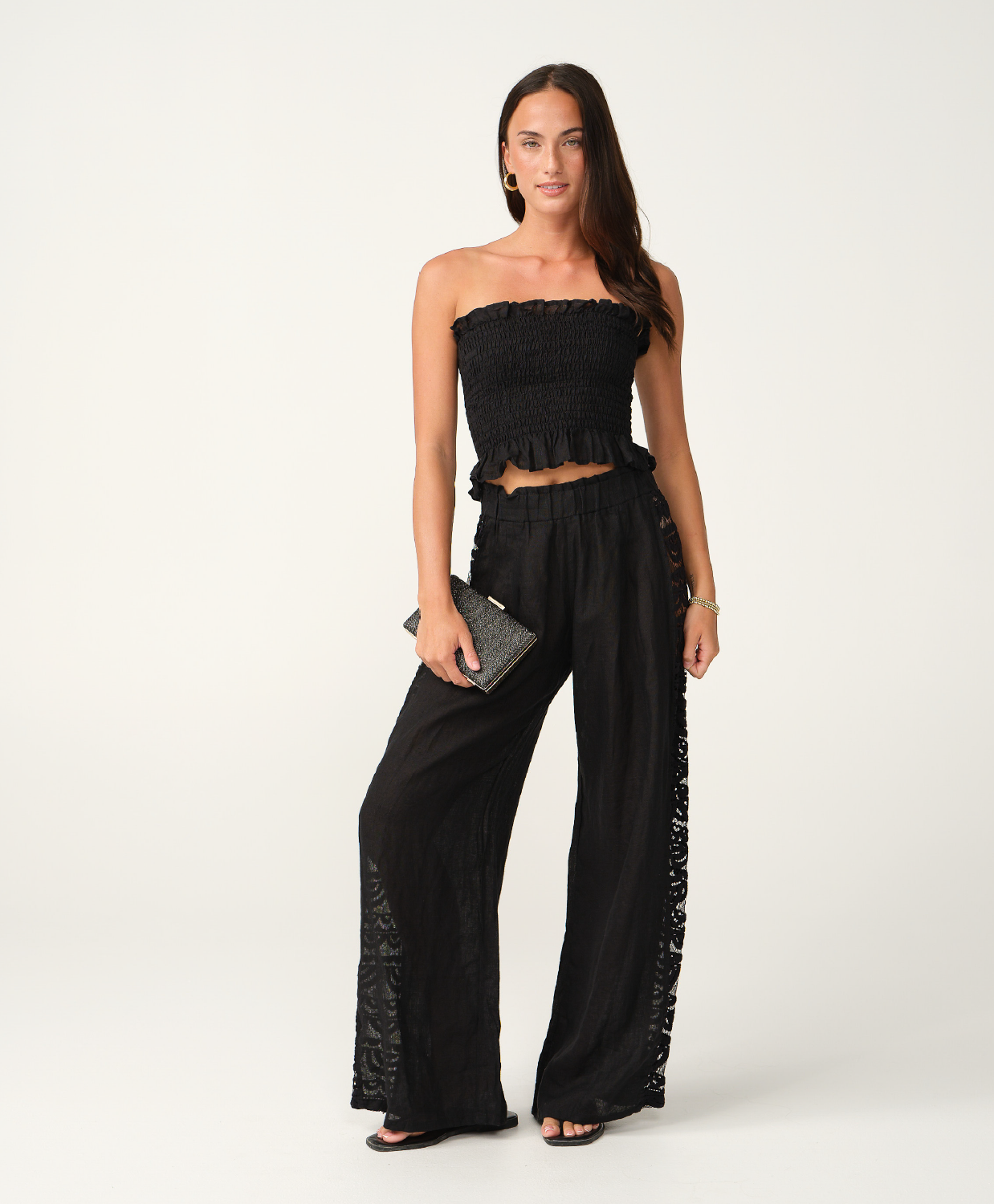 A woman modeling a black strapless cropped top and matching wide-leg pants with lace side panels, photographed against a clean off-white studio background.